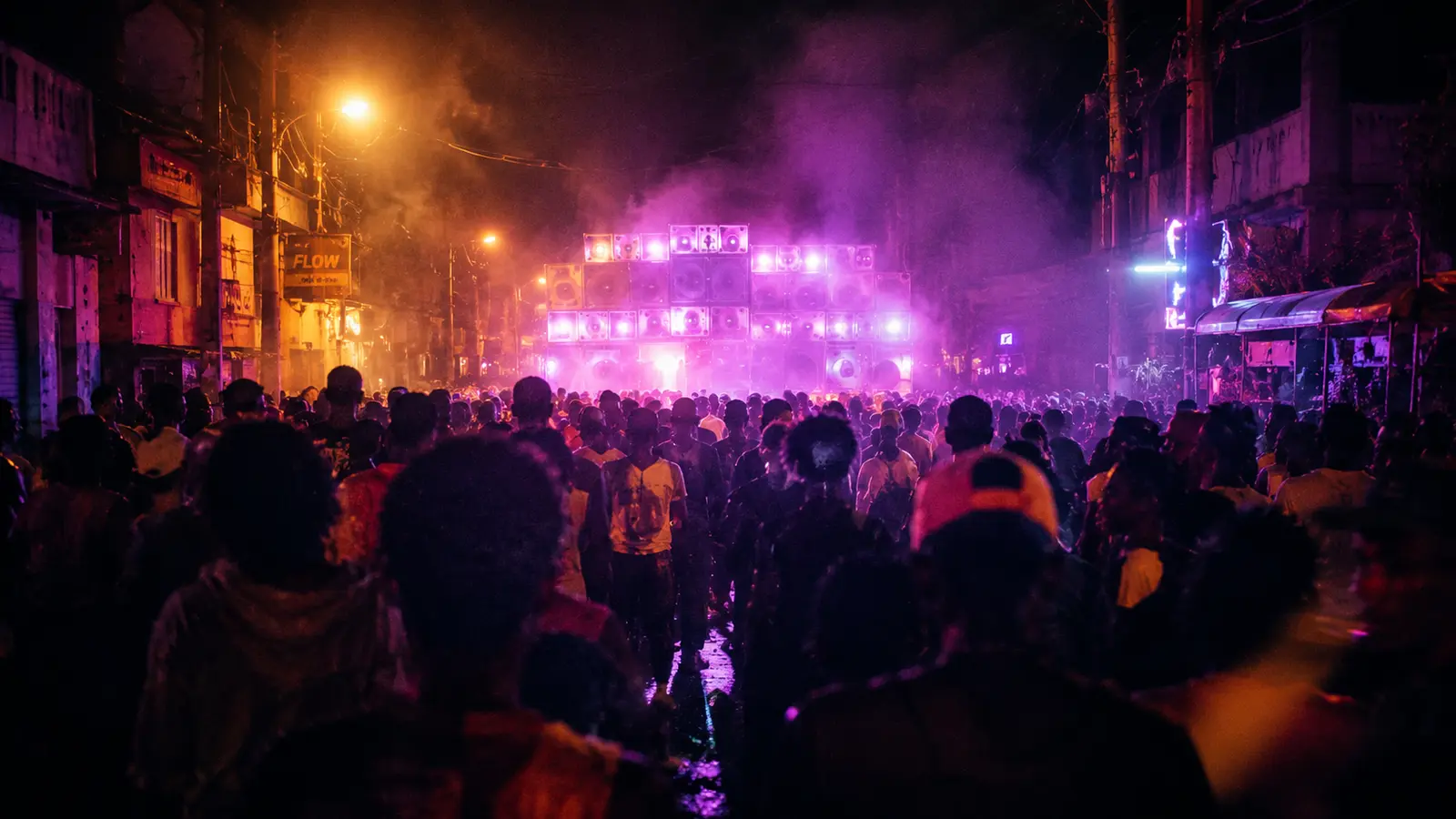 Foule de carnaval caribéen vue de dos en pleine nuit à Roseau La Dominique 1988-1989, sound system massif illuminé en arrière-plan, lumière tungstène + néons mauves, brouillard, palette magenta dominante
