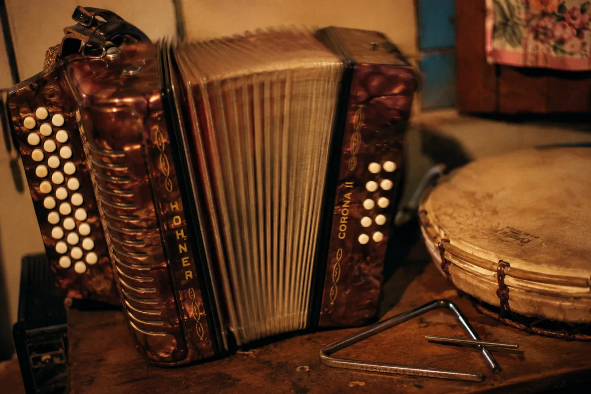 Vintage Hohner button accordion sitting on a wooden table, triangle and lapo kabwit beside it, Dominican interior 1988, tungsten light, slight motion on the bellows