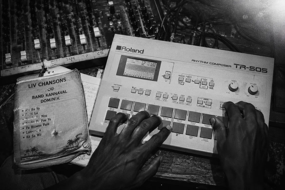 Roland TR-505 drum machine seen from above, hands programming a pattern, mixer cables around it, Creole lyric sheet, black and white Tri-X 1990