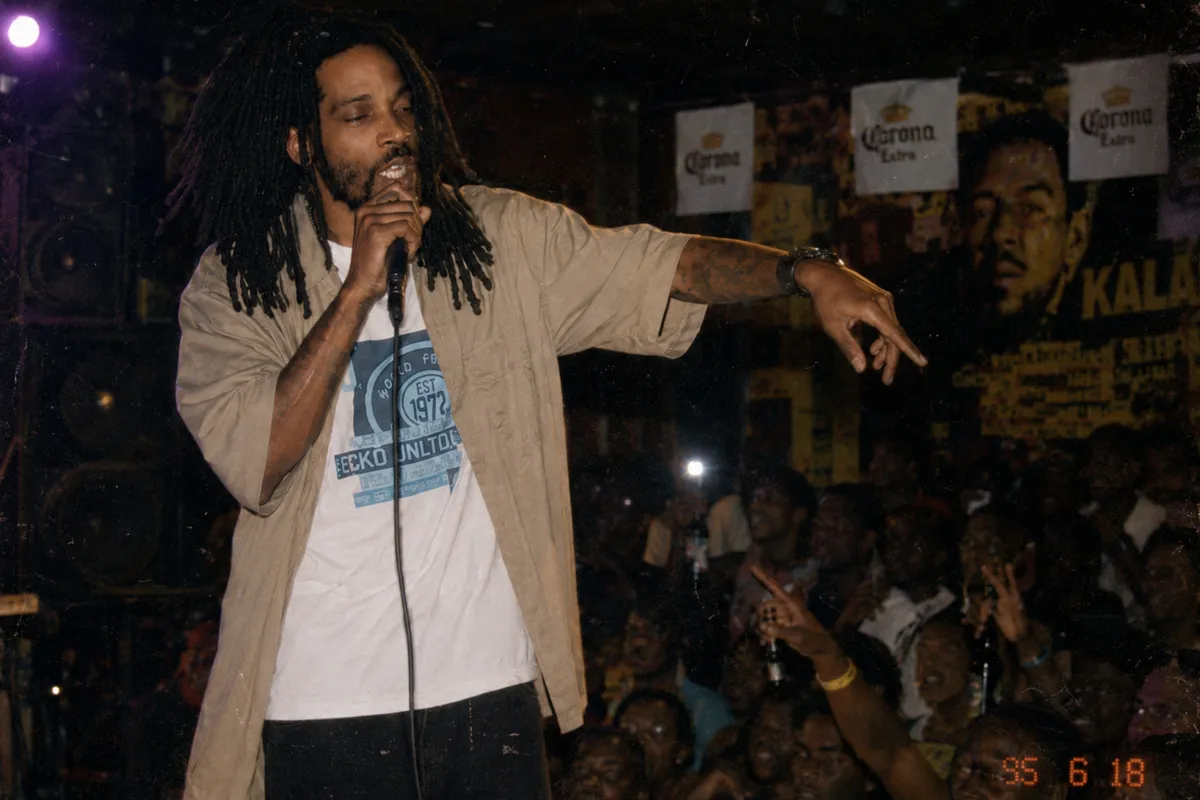 Young Caribbean singer in 1995 mid-phrase with mic in hand, sweat under the stage lights, PA stacks behind, dancehall + carnival ambience, tungsten light + magenta