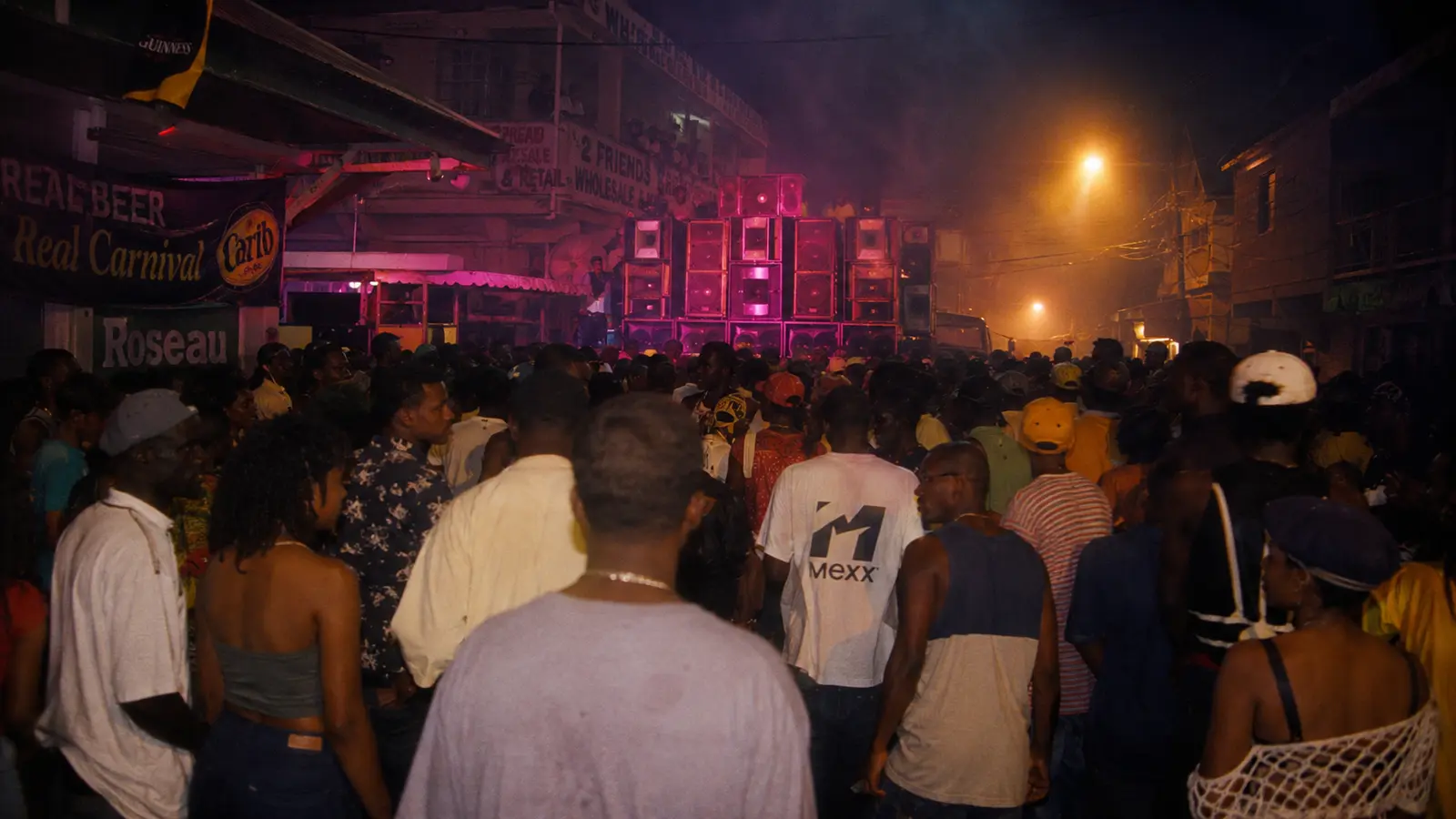 Caribbean carnival crowd seen from behind in the middle of the night in Roseau, Dominica 1996, massive illuminated sound system, tungsten + magenta neon, mist and humidity, asymmetric composition