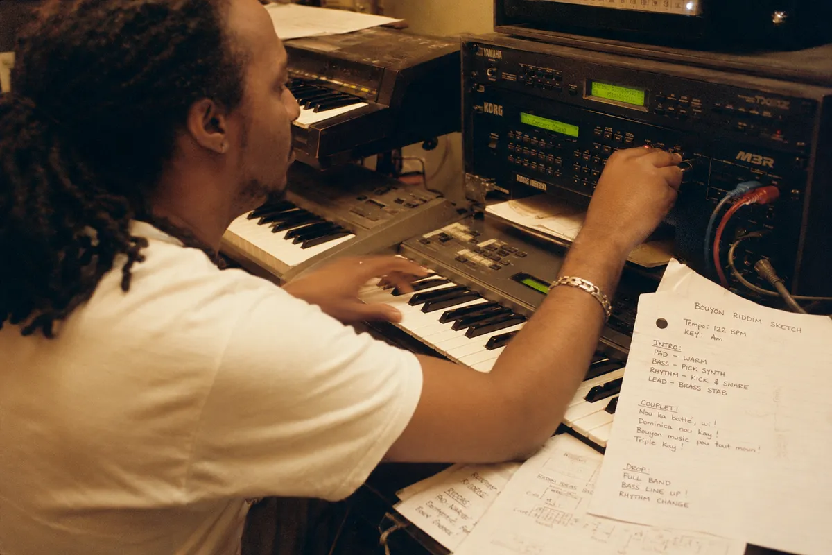 Vue par-dessus l'épaule d'un musicien caribéen programmant un rack de claviers en 2002, répétition Triple Kay à Roseau La Dominique, claviers vintage Yamaha ou Korg avec patch cables, mains en train de programmer, papiers avec esquisses de riddim Bouyon en créole, lumière tungstène d'en haut, léger flou de mouvement