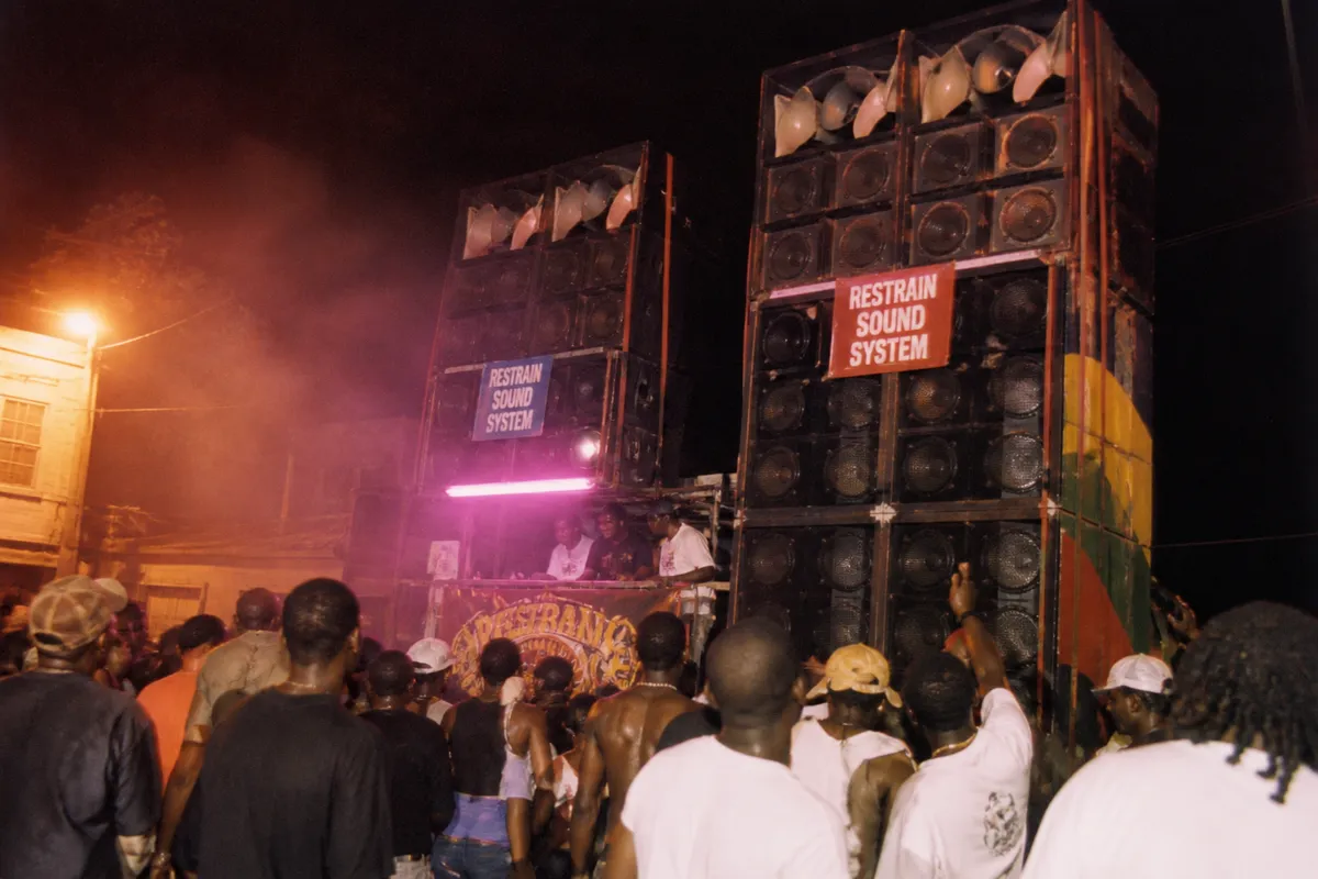 Plan large en contre-plongée d'un camion de sound system massif au carnaval de Roseau La Dominique vers 2005, double pile de baffles, foule dansant en avant-plan vue de dos, brume tungstène et néon magenta, poussière et humidité dans l'air, composition asymétrique