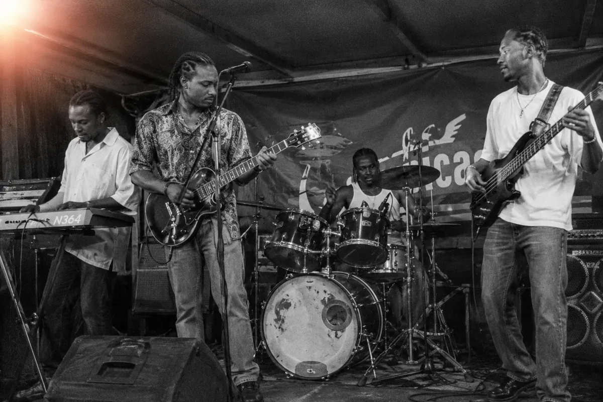 Plan moyen en noir et blanc d'un autre groupe Bouyon caribéen sur une scène de carnaval plus petite vers 2006, quatre musiciens visibles en pleine performance, batteur au centre, baffles PA plus modestes, ambiance de salle intime, fuite de lumière en haut à gauche, grain léger