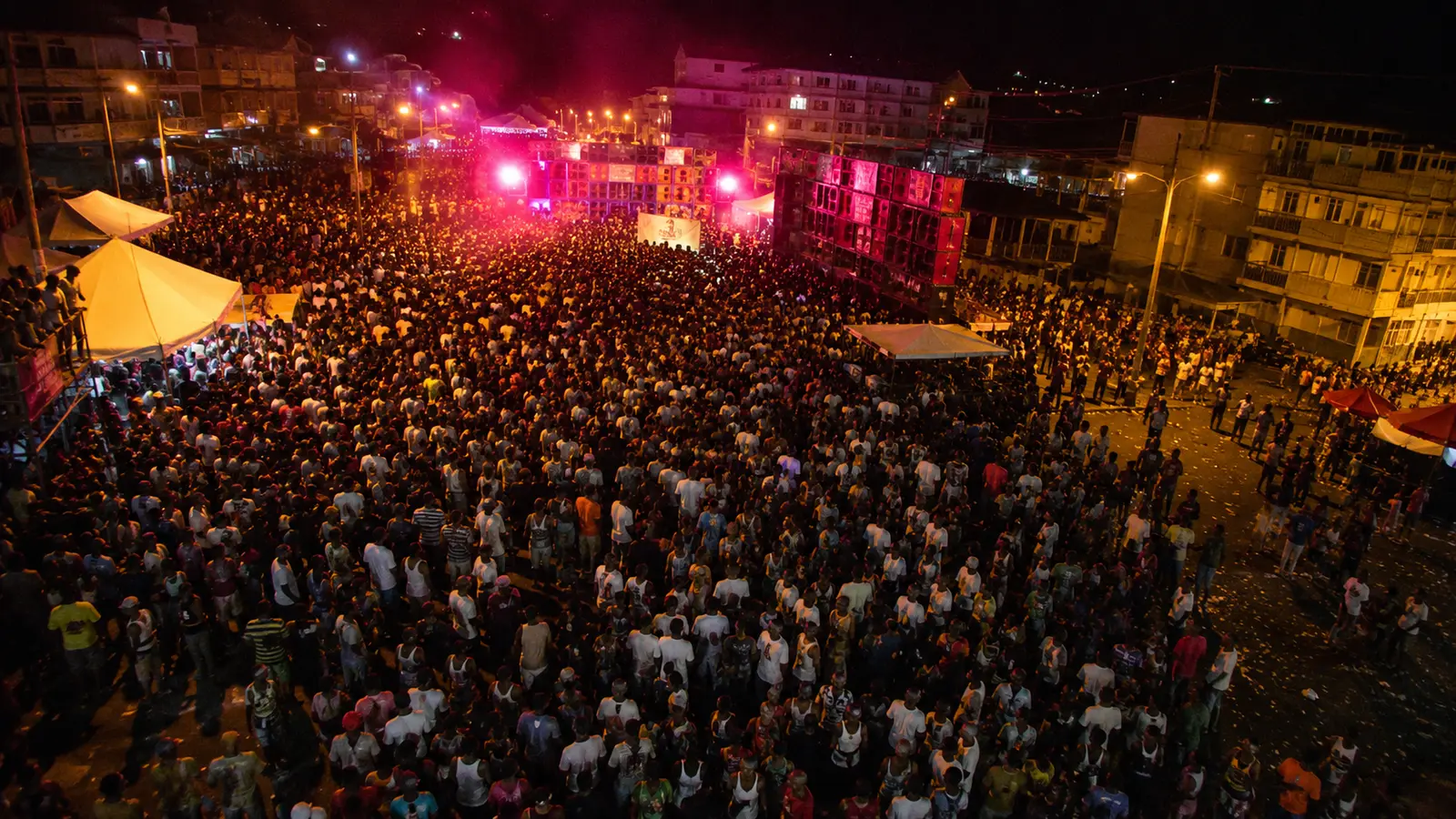 Plan très large d'une foule de carnaval caribéen vue d'en haut la nuit à Roseau La Dominique vers 2010, des milliers de personnes, piles de sound system illuminées en magenta et ambre, fumée et sueur, sensation de vide aux bordures avec quelques poches de foule plus clairsemées, mélange tungstène + néon magenta, composition asymétrique
