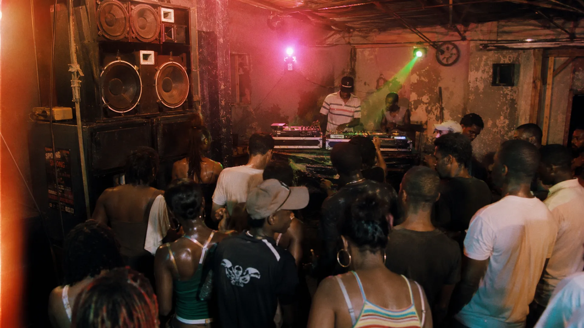 Wide shot of an underground Bouyon night in Pointe-à-Pitre Guadeloupe around 2009-2010, mobile sound system stacked left, mixing desk with two mixers, fifteen to twenty people dancing seen from behind, sweating, low ceiling, peeling paint walls, tungsten amber light + magenta neon haze + a single green strobe