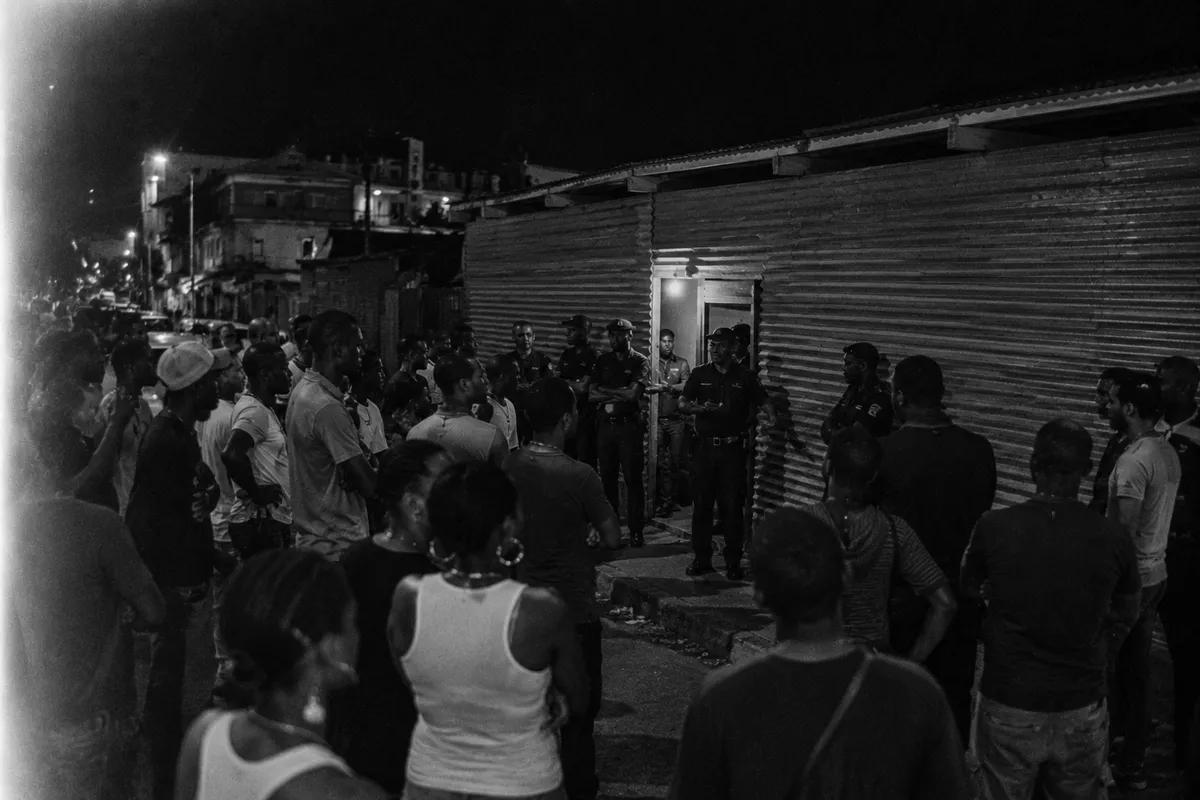 Wide shot of an empty Caribbean street at night around 2009 in Pointe-à-Pitre Guadeloupe, faint light from a closed venue door, police car parked at the corner with one officer visible, asymmetric framing, light leak top-left, visible grain, tense and hushed atmosphere