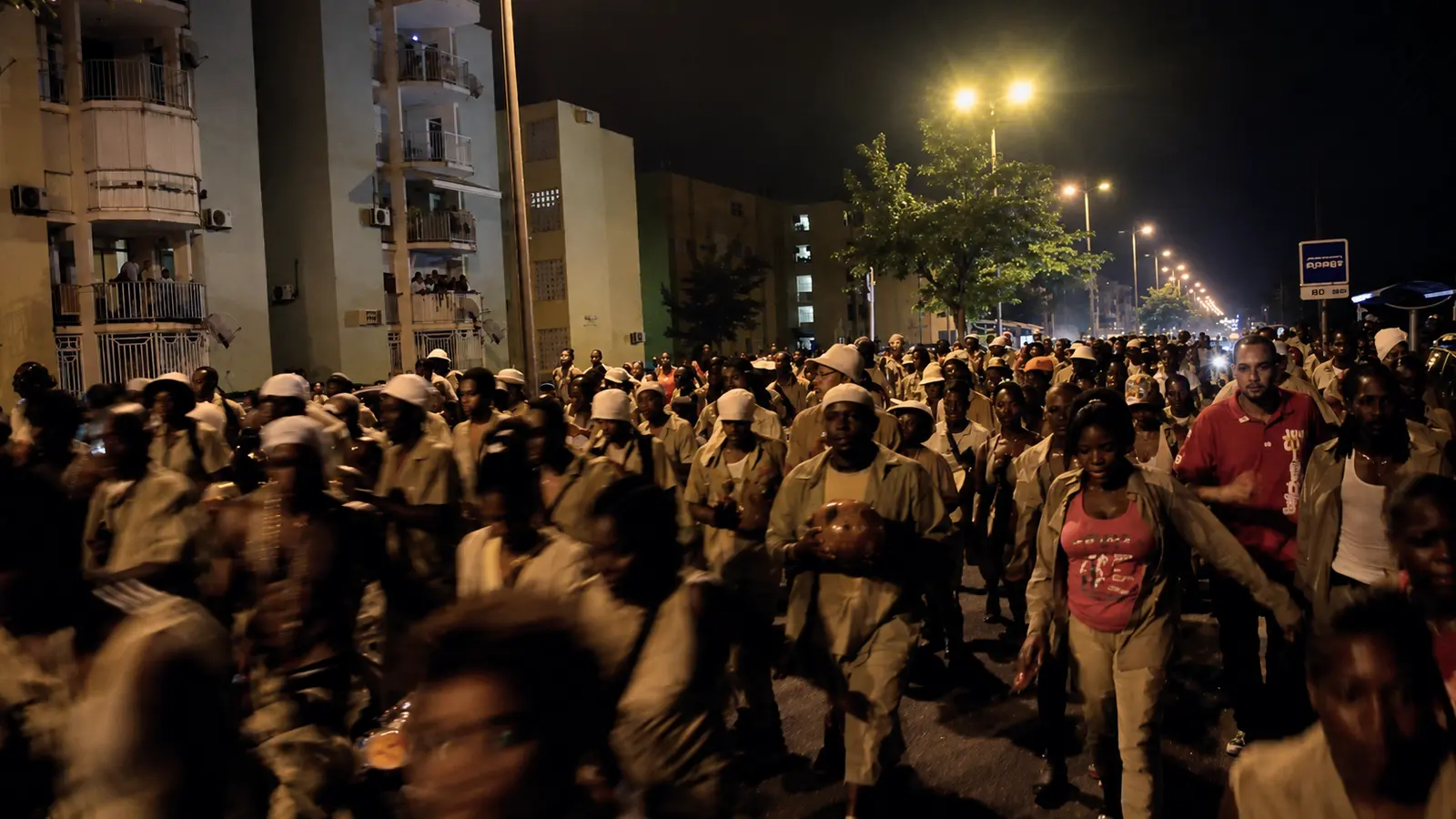 Very wide shot of a Caribbean carnival crowd at night in Pointe-à-Pitre Guadeloupe around 2013, brass band visible center, sound system in the distance, hundreds of people moving slowly through a narrow downtown street, magenta and amber tungsten haze mixing with deep blue night, smoke and humidity