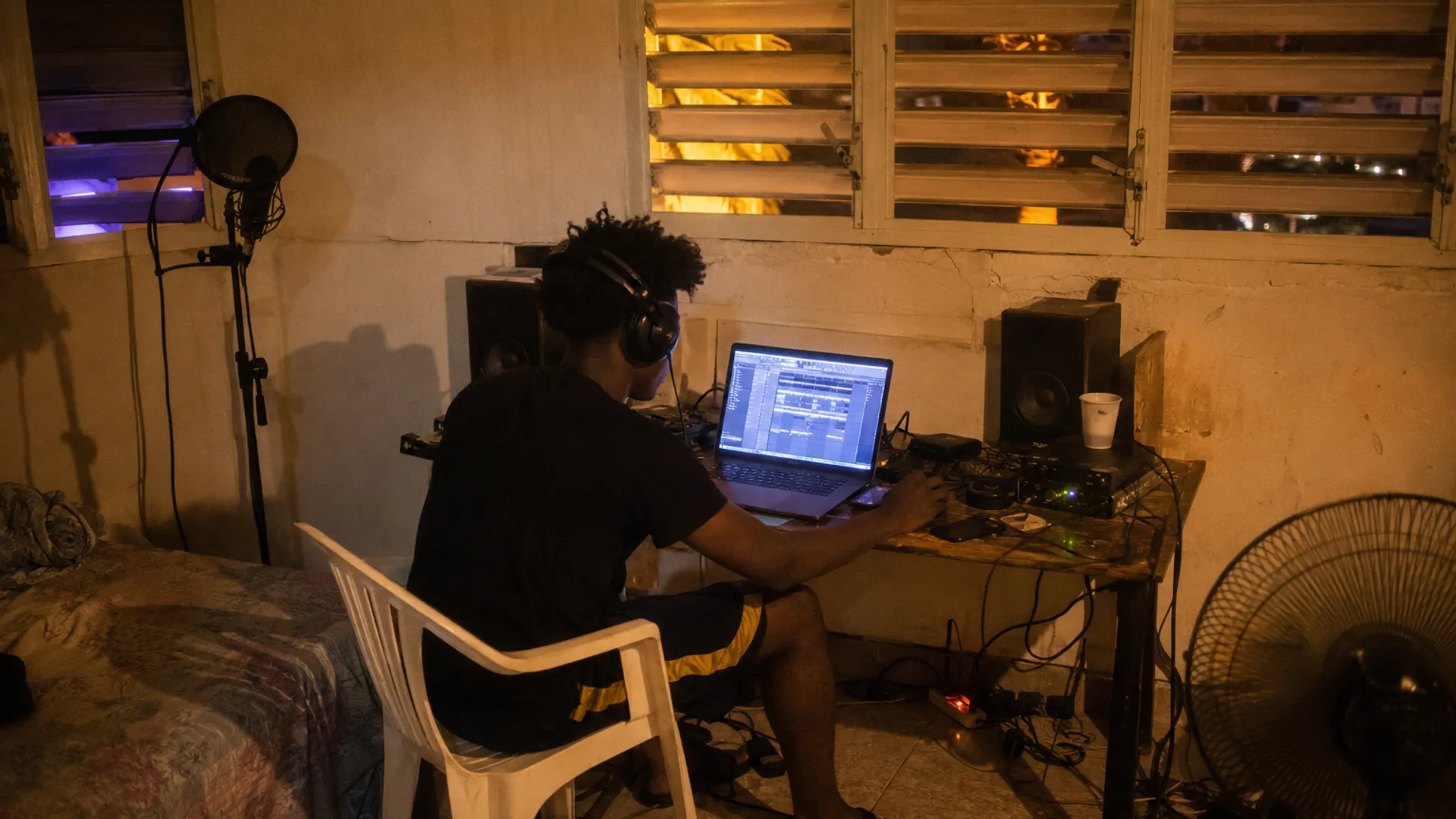 Petit studio maison guadeloupéen entre 2016 et 2022, ordinateur ouvert sur une session musicale sans texte lisible, interface audio, micro, câbles, clés USB, enceintes et chaise en plastique dans une chambre éclairée par une lumière chaude