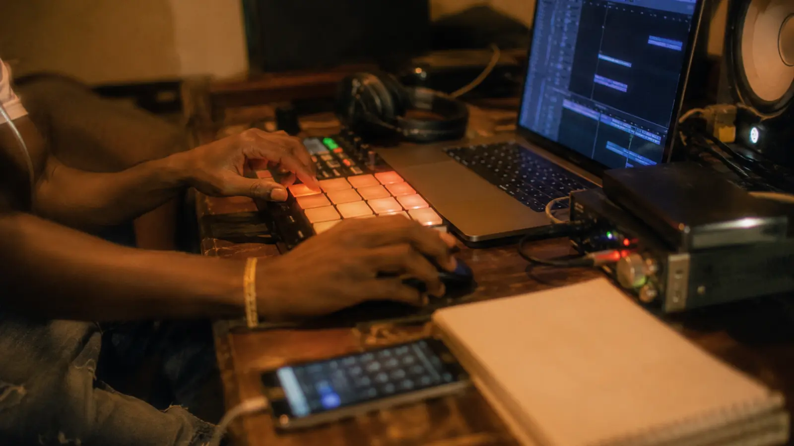 Gros plan réaliste sur des mains travaillant un beat pad et un ordinateur dans un petit studio guadeloupéen, avec casque, interface audio, câbles et carnet sans texte lisible