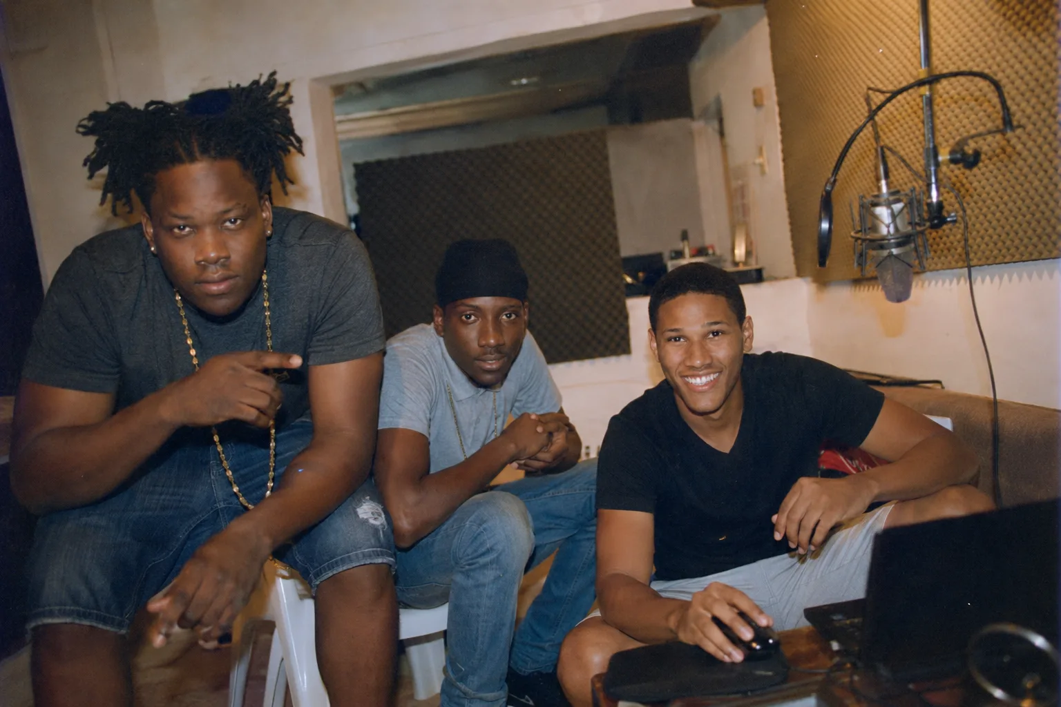 Trois jeunes artistes caribéens inspirés des Tolly Boyz dans un petit studio d'enregistrement à Saint-Martin en 2017, micro suspendu à droite, ordinateur portable au premier plan, murs avec panneaux acoustiques, lumière tungstène chaude, énergie concentrée de début de scène