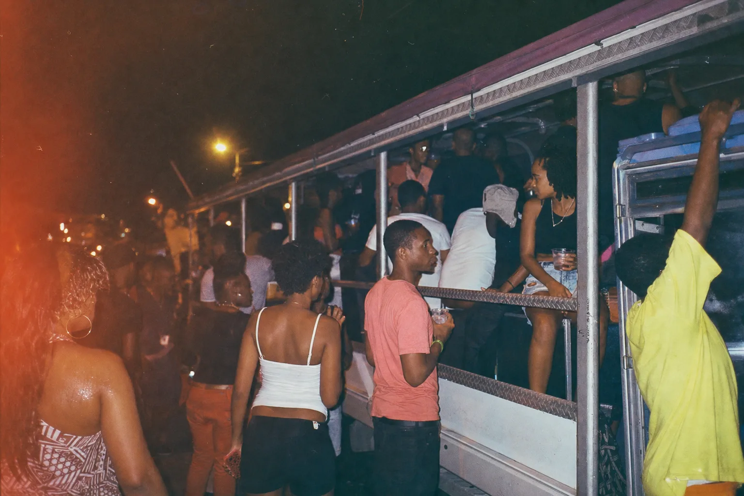 Scène de nuit à Sandy Ground Saint-Martin autour d'un party bus rempli de jeunes, ambiance humide, lumière tungstène, personnes avec gobelets, rue caribéenne animée avant une soirée