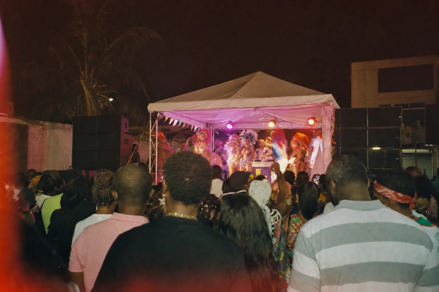 Foule vue de dos devant une petite scène de carnaval à Saint-Martin en 2017, chapiteau blanc, stacks de sound system, lumières magenta et ambre, costumes et téléphones levés