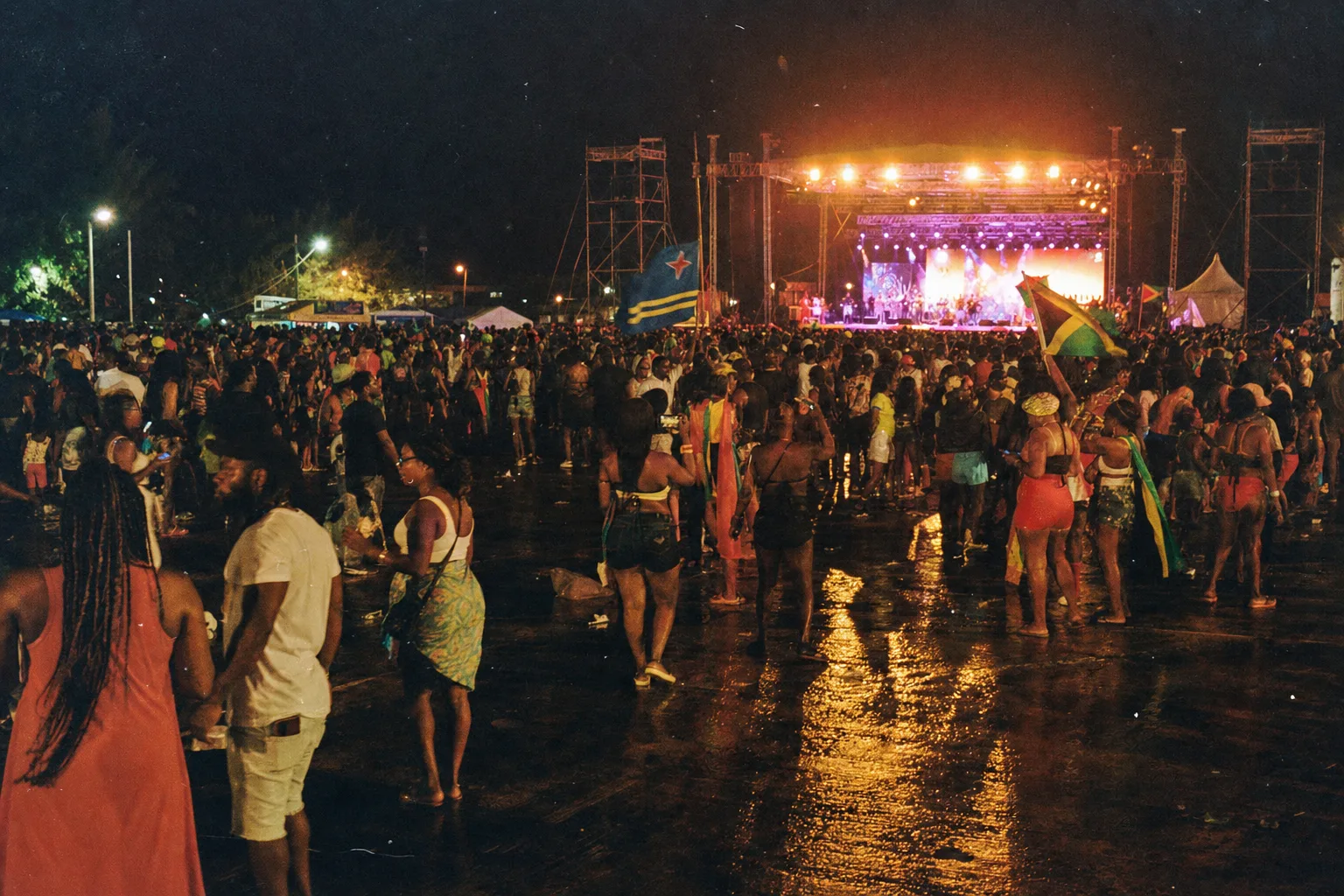 Grande foule de festival Bouyon à St. Maarten en 2025 devant une scène lumineuse, sol mouillé reflétant les projecteurs, drapeaux caribéens, sound system et public dense de nuit