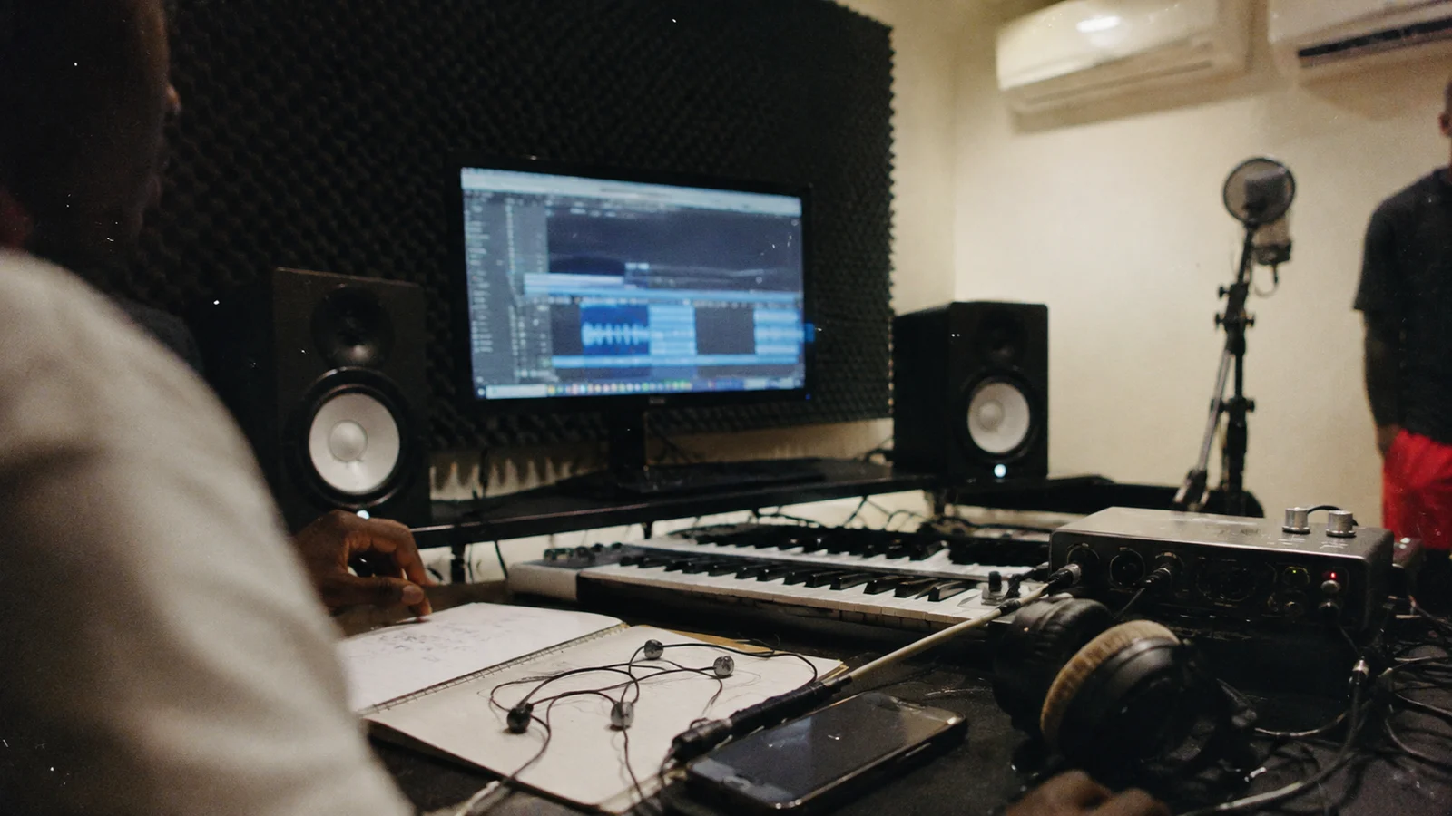 Documentary close-up of a music workstation with blurred audio screen, MIDI keyboard, earphones, audio interface, phone and cables in a Guadeloupean studio