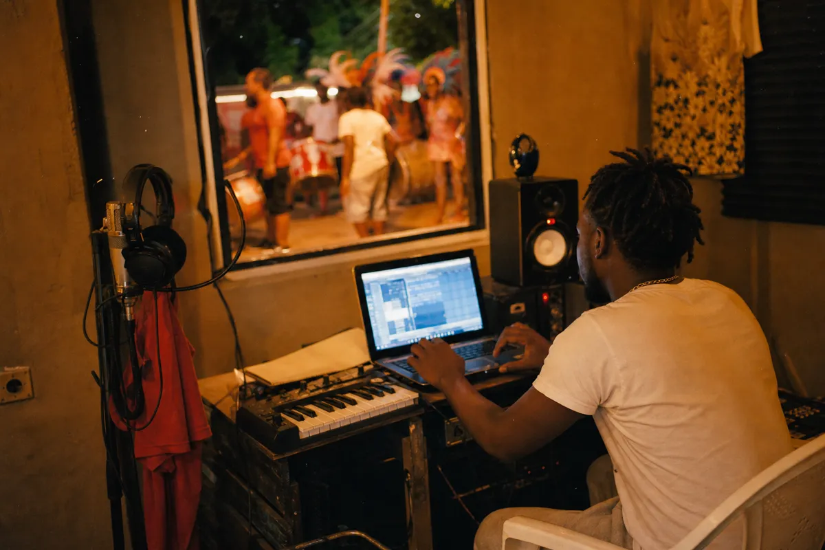 Modest Caribbean studio with producer, laptop, MIDI keyboard and blurred carnival energy outside