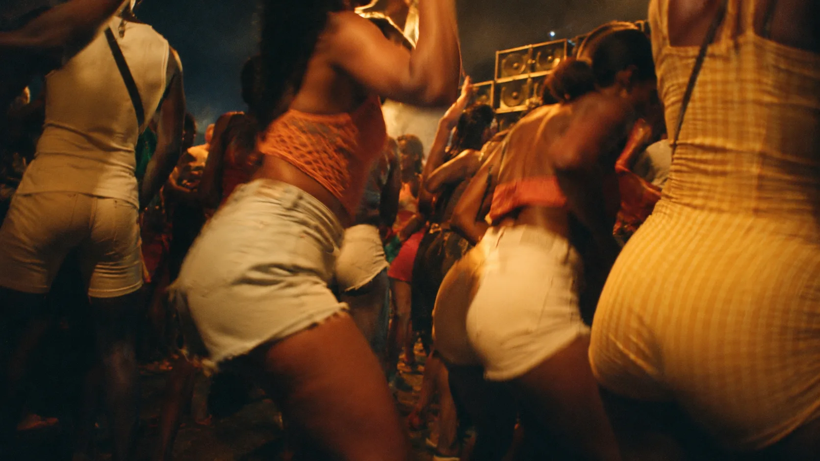 Crowd at a Caribbean night party dancing bouyon close together, hips and waist moving, a sound system in the background, festive energy.
